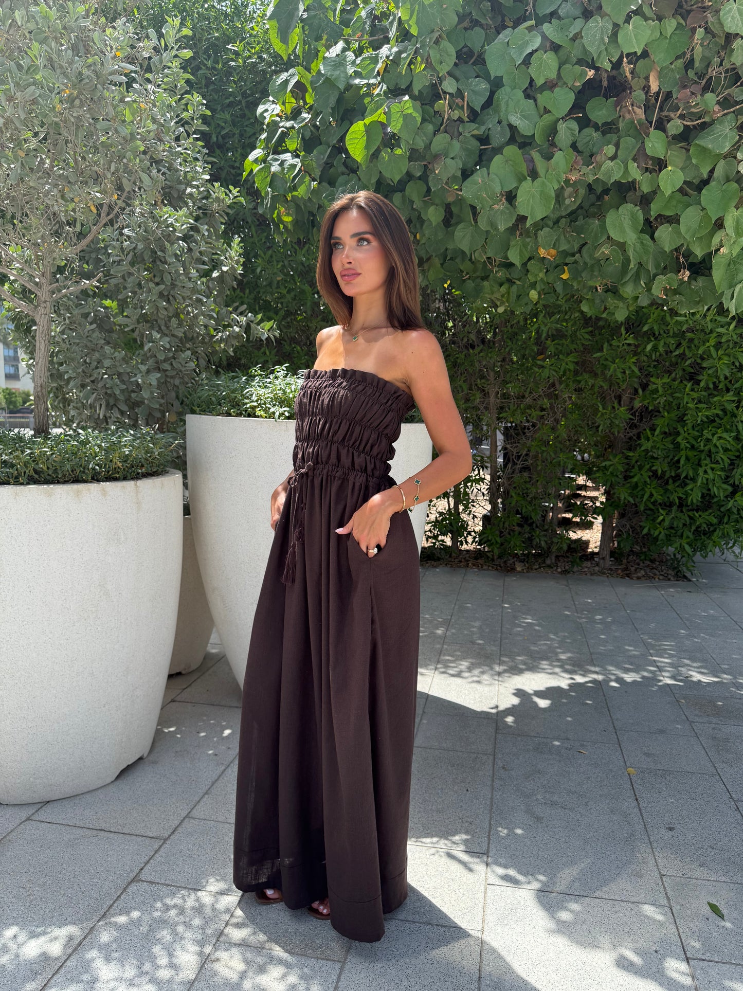 Woman wearing a strapless brown maxi dress with ruched bodice and wide-leg silhouette, standing outdoors in natural sunlight.