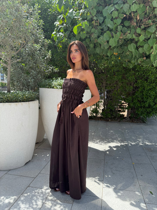 Woman wearing a strapless brown maxi dress with ruched bodice and wide-leg silhouette, standing outdoors in natural sunlight.